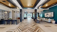 Located at 548 Lancaster Ave in Malvern, PA, this thoughtfully designed amenity space delivers a compelling blend of style and functionality across a single floor. The expansive lobby lounge immediately impresses with its striking herringbone-patterned tile flooring, coffered ceilings with warm gold accents, and dark structural columns that frame the open layout. Pendant lighting casts an inviting glow over multiple seating areas furnished with leather lounge chairs, contemporary sofas, and accent tables. A stone-clad, double-sided fireplace serves as a dramatic focal point, anchoring the social lounge area, while bold teal accent walls and curated artwork add vibrant character throughout. Large windows invite abundant natural light and views of the surrounding greenery. A dedicated leasing or reception area with a built-in counter and bar-height seating provides a professional yet welcoming point of contact. The conference room offers a refined setting for meetings, featuring a long wood-grain table, leather executive chairs, a wall-mounted display, and an elegant industrial-style chandelier overhead, with glass partition walls allowing visual connectivity. Completing the amenity offering is a state-of-the-art indoor golf simulator room, equipped with full simulation technology, turf flooring, and club storage — an exceptional recreational feature that sets this space apart.