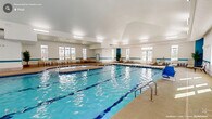 This generously sized indoor aquatic facility in Durham, NC, offers a well-appointed, single-level space designed for both recreational and fitness-oriented swimming. The centerpiece of the facility is a large rectangular lap pool with clearly marked swim lanes, complemented by an adjacent hot tub/whirlpool for relaxation. A broad pool deck surrounds the water on all sides, providing ample circulation space and furnished with sling-style lounge chairs and small tables arranged along the perimeter for comfortable poolside seating.

The architecture features vaulted ceilings that lend an open, airy feel to the interior. Rows of large, multi-pane windows line multiple walls, flooding the space with abundant natural light while offering views of the surrounding exterior. A teal accent band runs horizontally along the walls, adding a cohesive aquatic aesthetic to the otherwise crisp white interior. Flush and linear fluorescent lighting supplements natural illumination throughout.

Entry doors are positioned along the pool deck, and safety equipment, including a life ring, is visibly mounted near the pool edge. Potted tropical plants are strategically placed to enhance the ambiance. Overall, the facility presents a clean, functional, and welcoming environment suited for year-round aquatic use.