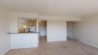 This single-floor unit offers a functional and straightforward layout designed for comfortable everyday living. The open-concept main area connects the living space to the kitchen, creating a cohesive and practical floor plan. The living area is finished with neutral-toned wall-to-wall carpeting, which extends throughout much of the unit, and benefits from natural light streaming through windows, casting bright patterns across the floor.

The kitchen is positioned along one side of the main living space, separated by a peninsula counter with a dark granite-style surface that provides additional workspace and casual seating potential. White cabinetry lines both the upper and lower sections of the kitchen, offering ample storage, while white appliances — including a refrigerator, electric range, and hood vent — contribute to the clean, cohesive aesthetic. A tile backsplash and light tile flooring complete the kitchen's practical design.

The bedroom is a generously sized, carpeted room with a window that frames outdoor greenery, filling the space with natural light. A walk-in closet provides substantial storage, with an additional door leading to the bathroom. The bathroom features a full tub-shower combination with classic white subway tile, a granite-topped vanity, and tile flooring, offering a clean and functional design throughout.
