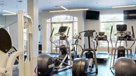 Located at 1 Richmond St in New Brunswick, NJ, this modern fitness center offers a thoughtfully designed, single-floor layout divided into distinct workout zones. The main cardio area features an array of Precor ellipticals, upright and recumbent bikes, and treadmills, all positioned near large arched windows that flood the space with natural light and provide views of the surrounding streetscape. Wall-mounted televisions add an entertainment element for cardio sessions. The strength training zone is expansive and open, lined with floor-to-ceiling mirrors that amplify the sense of scale. White Cybex cable machines and multi-station weight equipment are arranged across rubber flooring, complemented by a dramatic cityscape panoramic mural that serves as a striking focal point. A dedicated functional fitness studio features light hardwood-style flooring, a slatted wood ceiling accent, full-length mirrors, a wall-mounted digital training screen, medicine ball storage, and open floor space ideal for group classes or personal training. The facility also includes a cable crossover station near the main entrance, bathed in natural light from arched glass doors. Throughout, linear pendant lighting, white columns, and a clean neutral palette create a bright, professional, and motivating environment.