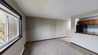 Located at 5150 16th Ave SW in Cedar Rapids, Iowa, this single-floor unit offers a practical and comfortable layout designed for everyday living. The open living area is generously sized, finished with neutral gray walls, plush carpeting, and baseboard heating, creating a clean and inviting atmosphere throughout.

The kitchen is efficiently arranged and features dark wood cabinetry, granite-style countertops, black appliances including an electric range, over-the-range microwave, and refrigerator, along with a sink. A pass-through opening connects the kitchen to the living area, enhancing the open feel of the space. The kitchen is finished with light wood-look flooring.

The bedroom is a comfortable, well-proportioned room equipped with a ceiling fan, a large window that brings in natural light and views of the surrounding greenery, and a wall-mounted air conditioning unit alongside baseboard heating for year-round climate control. Soft carpeting continues throughout the bedroom.

The bathroom is accessed through a dark-trimmed door and features light wood-look tile flooring, a dark wood vanity with a light countertop, a toilet, and a tub and shower combination enclosed by sliding frosted glass doors, accented by soft blue walls. The unit also includes an entry area with a classic six-panel door. Overall, the space presents a functional, move-in-ready design.