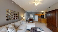 Located at 111 Orchard Hills Dr in Jeffersonville, Indiana, this single-floor residence offers a well-organized, open-concept layout designed for comfortable everyday living. The living room is a welcoming focal point, featuring vaulted ceilings with a ceiling fan, plush carpeting, and a large sliding glass door fitted with vertical blinds that fill the space with natural light and provide access to the outdoors. The area flows seamlessly into a dedicated dining space, illuminated by a hanging chandelier, and then into the kitchen. The kitchen is equipped with warm oak cabinetry, granite-style countertops, a breakfast bar peninsula, tile flooring, and standard appliances including a refrigerator, electric range, and built-in microwave. Two well-proportioned bedrooms each feature carpeted floors, large windows with outdoor views, and ample natural light. Both bathrooms include warm wood vanities with white countertops and tile flooring; one bathroom features a standalone walk-in shower with a framed glass door. A dedicated utility room houses the water heater and washer/dryer hookups, adding practical convenience. The consistent use of neutral gray tones throughout the space creates a cohesive and modern ambiance.