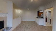 This single-floor residence offers a clean, open layout designed for comfortable everyday living. The spacious main living area is finished with plush wall-to-wall carpeting and crisp white walls accented by elegant crown molding, creating a bright and welcoming atmosphere. Recessed lighting and a flush-mount ceiling fixture provide ample illumination throughout the space, while a ceiling fan adds to the comfort of the room. A half-wall partition subtly defines the transition between the living area and the adjoining kitchen, maintaining an open and airy feel. The kitchen is thoughtfully appointed with warm wood cabinetry, a built-in microwave, and a dark stone countertop that extends to form a convenient breakfast bar — ideal for casual dining or meal prep. White appliances and tiled backsplash details add a clean, functional aesthetic to the cooking space. Neutral tones throughout the home create a versatile canvas, making it easy to personalize. The overall ambiance is calm and move-in ready, with a layout that balances openness with defined functional zones.