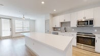 Located in Brookshire, TX, this single-story, one-bedroom, one-bathroom unit offers a thoughtfully designed open-concept layout that maximizes its compact footprint. The kitchen serves as a central highlight, featuring crisp white shaker cabinetry with matte black hardware, a grey subway tile backsplash, stainless steel appliances including a gas range, microwave, and refrigerator, and a generous island with white quartz countertops providing ample workspace and storage. A built-in desk nook adjacent to the kitchen adds functional versatility. Light wood-look flooring flows throughout the main living areas, complementing the bright, neutral palette. The living room receives abundant natural light through large windows and a glass-paneled door, with a ceiling fan overhead. The bedroom is carpeted and similarly well-lit, offering a comfortable, quiet retreat with a ceiling fan and dedicated closet. The bathroom is finished with large-format white wall tiles surrounding the tub-shower combination, accented by matte black fixtures, a white vanity, and matching wood-look flooring. Throughout the unit, recessed lighting and a clean, contemporary aesthetic create a cohesive and inviting atmosphere.