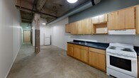 Located in the heart of Saint Paul, MN, Unit 308 at 281 5th Street East is a compact yet characterful industrial loft that embraces its raw architectural roots. The unit features polished concrete floors throughout, exposed concrete columns, and open ductwork and piping along the ceiling, all of which contribute to its distinctive urban loft aesthetic.

The main living area is an open, flexible space defined by crisp white walls that contrast with the industrial structural elements. A built-in kitchen lines one wall, outfitted with light maple cabinetry, dark countertops, a double-basin sink, and space for appliances including a refrigerator and range. Overhead pendant lighting adds warmth to the functional kitchen area.

A split-level layout adds architectural interest, with a staircase and black metal railing connecting two distinct floor levels within the unit. The upper level benefits from natural light filtering in through windows, offering views to the exterior.

The bathroom is clean and efficiently designed, featuring a toilet, sink, and grab bars, with a caged wall sconce providing ambient lighting. A dedicated closet provides convenient storage. A long hallway connects the various areas of the unit, enhanced by a natural wood door element that adds warmth against the otherwise industrial palette. The overall ambiance is minimalist, urban, and full of character.