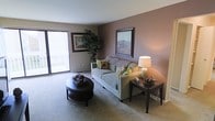 This single-floor space offers a thoughtfully designed living environment with a focus on comfort and style. The living room serves as the heart of the space, featuring a warm mauve accent wall that adds depth and character to the room. Soft, neutral carpeting runs throughout, contributing to the cozy and inviting ambiance. A large sliding glass door floods the space with an abundance of natural light while offering views to the outside, creating an airy and open feel. The room comfortably accommodates a plush sofa adorned with decorative pillows, a classic round coffee table, and coordinating end tables, all arranged to encourage relaxed conversation and everyday living. Framed landscape artwork is displayed on the walls, adding a refined, curated touch to the interior. A lush indoor plant nestled near the window brings a natural, organic element into the space. The overall design aesthetic balances understated elegance with practicality, making it a welcoming retreat. Neutral tones, warm lighting, and clean lines work together to create a harmonious living environment that feels both functional and inviting.