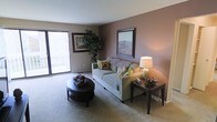 This single-floor space offers a thoughtfully designed living environment with a focus on comfort and style. The living room serves as the heart of the space, featuring a warm mauve accent wall that adds depth and character to the room. Soft, neutral carpeting runs throughout, contributing to the cozy and inviting ambiance. A large sliding glass door floods the space with an abundance of natural light while offering views to the outside, creating an airy and open feel. The room comfortably accommodates a plush sofa adorned with decorative pillows, a classic round coffee table, and coordinating end tables, all arranged to encourage relaxed conversation and everyday living. Framed landscape artwork is displayed on the walls, adding a refined, curated touch to the interior. A lush indoor plant nestled near the window brings a natural, organic element into the space. The overall design aesthetic balances understated elegance with practicality, making it a welcoming retreat. Neutral tones, warm lighting, and clean lines work together to create a harmonious living environment that feels both functional and inviting.
