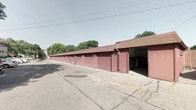 This single-story facility features a continuous row of individual garage units, each finished in a consistent mauve brick-red tone that gives the complex a cohesive, well-maintained appearance. The units are constructed with concrete block walls and topped with a matching red-brown shingled roofline, lending the structure a sturdy, durable character. Each unit is equipped with its own overhead roll-up door, providing private, enclosed access for vehicles, equipment, or storage needs. A wide, paved concrete apron runs the full length of the building, offering ample room for vehicle maneuvering and straightforward drive-up entry. A downspout is integrated into the façade for efficient water drainage. The surrounding environment features open sky and mature trees in the background, giving the facility a relatively open and accessible setting. The layout is straightforward and functional, with each unit separated by shared walls, maximizing the number of individual spaces within the row. The overall design prioritizes practicality and ease of use, making this facility well-suited for secure vehicle storage, light utility purposes, or additional property storage needs. The uniform exterior finish and organized row configuration contribute to a tidy, orderly presentation of the space.