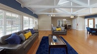Located at 3831 Brentwood Rd in Raleigh, NC, this single-story space makes a striking first impression with its dramatic vaulted ceilings featuring exposed white painted beam trusses that run the length of the main area, creating an airy, open atmosphere. Warm hardwood flooring flows throughout, unifying the various functional zones seamlessly.

The expansive main area is bathed in natural light from large windows fitted with white plantation shutters, offering views of the surrounding greenery. This open layout accommodates a comfortable lounge area with leather sofas accented by navy and yellow throw pillows, anchored by a plush navy area rug and a glass-top coffee table — ideal for relaxed gatherings or meetings.

A dedicated office area features a substantial dark wood executive desk with ample workspace, complemented by a matching filing cabinet and a decorative console table topped with a wall-mounted mirror. Ceiling fans with chandelier-style fixtures provide both function and elegance throughout the space.

Toward the rear, a dining area is furnished with a glass-top table and dark wood chairs beneath a chandelier, with framed artwork adorning the shiplap-accented walls. A wood-framed glass door leads to the exterior, allowing additional light to fill the space. The overall design blends a welcoming residential warmth with professional functionality.
