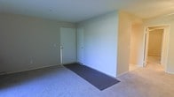 This single-floor residential space presents a straightforward, functional layout anchored by a carpeted main living area. The entryway is defined by a standard white door equipped with a brass-toned handle and deadbolt, set against smooth, neutral-toned walls that lend the space a clean, understated appearance. A rectangular runner mat sits at the entry transition point, providing a practical surface between the door and the main carpeted floor. The wall-to-wall carpet throughout the living area is a light gray tone, contributing to the overall neutral aesthetic of the space. The walls are painted in a soft, warm beige, creating a simple and cohesive color scheme. A partial wall or divider is visible to the right side of the room, suggesting a connected but defined adjacent space, likely a hallway or secondary room, with a similarly warm wall tone. Baseboard heating units are visible along the lower wall, indicating the building's primary heating method. The ceiling is smooth and standard height, and the overall layout appears open and easy to configure for a variety of uses.