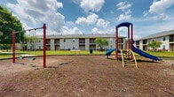 Located at 1900 Cardinal Trail Dr in Galloway, Ohio, this outdoor community space offers a welcoming recreational area for residents. The grounds are open and expansive, providing plenty of room for outdoor activities and social gatherings. A standout feature of this space is the colorful playground structure, equipped with slides, climbing ladders, and vertical poles, making it an engaging destination for younger residents and families. The playground is set on a dirt and mulch surface, designed to provide a safe play environment. A picnic bench is positioned nearby, offering a convenient spot for supervising adults or those looking to enjoy the outdoors. The surrounding lawn is green and well-maintained, bordered by a multi-story residential building featuring balconies and neutral-toned exterior siding. Mature trees dot the landscape, adding shade and a natural aesthetic to the area. The open-sky setting allows for abundant natural light throughout the day, creating a bright and inviting atmosphere. This community outdoor space combines functional recreational amenities with a comfortable, accessible layout suited for everyday use.