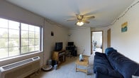 This single-floor space centers around a comfortable living room that sets a casual, functional tone throughout. The living room features wall-to-wall carpeting that adds warmth underfoot, and a ceiling fan with an integrated light fixture that provides both airflow and ambient lighting. A wall-unit air conditioner is installed beneath one of the windows, ensuring year-round comfort. Natural light enters through the windows, brightening the neutral-toned walls and open layout.

The living area is thoughtfully arranged to accommodate both relaxation and entertainment, with space for a sofa, coffee table, television console, and a dedicated desk area. String lights are draped along the upper perimeter of the walls, adding a decorative accent to the room. A floor lamp in the corner provides additional task lighting.

A doorway visible from the living room leads into an adjoining room, suggesting an open and connected floor plan that flows naturally between spaces. The overall ambiance of the space is relaxed and lived-in, with a practical layout suited for everyday living. The neutral color palette, carpeted floors, and modest finishes give the space a straightforward, welcoming character that is easy to personalize and arrange.