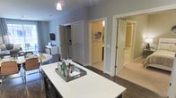 This single-floor residence features an open-concept layout that seamlessly connects the kitchen and living areas. The kitchen is anchored by a generous white quartz-topped island with seating, providing both a functional workspace and a casual dining option. Dark hardwood flooring runs through the main living areas, creating a striking contrast against the light gray walls and white trim throughout. A pendant light fixture hangs above, adding a focused point of illumination to the space. The living area beyond the island is carpeted and includes a media console and soft area rug, contributing to a comfortable, relaxed atmosphere. Multiple doorways visible from the main space lead to additional rooms, including what appears to be a bathroom with tiled walls and a separate room with carpeted flooring, likely a bedroom. A closet with louvered doors adds practical storage. The neutral color palette, clean lines, and modern finishes give the space a contemporary feel. Natural light filters through window treatments in the living area, enhancing the open and airy quality of the interior.