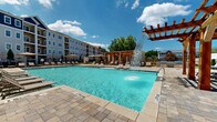 Located at 1875 Maple Dr in Kennesaw, Georgia, this outdoor amenity area offers a well-appointed, resort-style experience designed for relaxation and recreation. The centerpiece is a generously sized swimming pool with crystal-clear turquoise water, highlighted by a dramatic water feature that cascades from a striking wooden pergola structure. The pool deck is finished with elegant stone pavers that extend throughout the entire outdoor space, lending a cohesive and upscale aesthetic.

Surrounding the pool, multiple teak-framed lounge chairs with neutral sling seating are thoughtfully arranged to maximize sun exposure and comfort. Additional pergola structures provide shaded retreats along the perimeter, where lounge seating offers a cooler alternative for warm Georgia days. A covered patio area adjacent to the main building features ceiling fans, a ping-pong table, and direct access to the interior via large glass double doors, seamlessly blending indoor and outdoor living.

The building facade incorporates warm-toned brick masonry paired with crisp white board-and-batten siding, contributing to a modern yet inviting architectural character. Iron fencing and gated access points define the boundaries of the space while maintaining an open, airy feel. Mature trees and landscaping along the edges provide natural privacy and greenery, completing this thoughtfully designed outdoor retreat.
