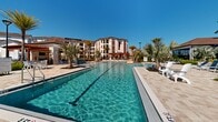 This two-level resort-style amenity complex offers an impressive outdoor experience centered around two generously sized swimming pools set within a beautifully designed paver courtyard. The pools feature crystal-clear water and are complemented by a dedicated lap lane, making them suitable for both leisurely recreation and active swimming. Surrounding the pools, wide stone-paved decks provide ample space for lounging, furnished with chaise lounge chairs and seating areas ideal for relaxation under the open sky.

Tropical landscaping throughout the space includes mature palm trees, fan palms, ornamental shrubs, and layered plantings that lend a lush, resort-like atmosphere. The grounds are framed by a well-maintained clubhouse building featuring white columns, a covered pergola area, and large glass entry doors, offering a welcoming architectural focal point. An additional covered pavilion structure provides shaded gathering space adjacent to the pool deck.

The overall ambiance is bright, open, and sun-drenched, consistent with a warm Florida climate setting. Multi-story residential buildings with distinctive red and neutral façades rise in the background, contributing to the sense of a cohesive, well-planned community. Thoughtful outdoor lighting and clean, modern hardscaping complete the polished aesthetic of this expansive outdoor amenity area.