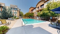 Located at 914 W Lake St in Fort Collins, Colorado, this outdoor amenity area serves as a well-appointed communal retreat for residents. The centerpiece is a generously sized rectangular swimming pool with clear, blue-tinted water, surrounded by a broad concrete pool deck that offers ample space for sunbathing and relaxation. Multiple adjustable lounge chairs and seating arrangements are positioned poolside, providing comfortable spots to enjoy Colorado's abundant sunshine.

The pool area is enclosed by a wrought iron security fence, ensuring a safe and private environment. Mature deciduous and evergreen trees frame the space, offering natural shade and a lush, park-like atmosphere. Ornamental grasses and low-maintenance plantings add texture and color throughout the landscaped grounds.

Adjacent to the pool, a paved courtyard patio features additional outdoor seating, including cushioned chairs and side tables arranged for casual gatherings. This secondary lounge area is shaded by large, established trees and connects seamlessly to the surrounding multi-story residential buildings, which feature private balconies overlooking the courtyard below.

The overall ambiance is that of a well-maintained, resort-style outdoor space designed to encourage community and outdoor living, set within the vibrant neighborhood of Fort Collins, CO.