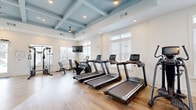 Located at 3625 Frankford Ave in Panama City, FL, this fitness center offers a thoughtfully designed space for a comprehensive workout experience. The room is immediately distinguished by its striking coffered ceiling, painted in a soft coastal blue and accented with recessed lighting and pendant fixtures, lending the space a polished yet welcoming ambiance.

Light wood-style flooring runs throughout, complementing the bright white walls and creating a clean, modern aesthetic. Multiple windows fitted with white plantation shutters line the walls, allowing generous natural light to fill the space while providing views of the surrounding outdoor area, including a pool visible through the glass-paneled exit doors.

The fitness center is well-stocked with a diverse selection of equipment. Cardio offerings include multiple treadmills and a stationary bike, each equipped with individual display consoles. Strength training is supported by a cable machine, multi-station weight machine, adjustable bench, a full dumbbell rack, and a medicine ball storage tower. A large wall mirror enhances the sense of openness and supports proper form during workouts. A wall-mounted flat-screen television adds to the functionality of the space. The layout provides ample open floor space, ensuring easy movement between stations and accommodating a variety of exercise routines.