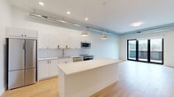 This single-floor unit showcases a contemporary industrial-modern design throughout. The open-concept kitchen and living area serves as the heart of the space, featuring flat-front white cabinetry, a white subway tile backsplash, stainless steel appliances including a built-in microwave and range, and a large central island with a clean white countertop. Pendant lights hang above the island, while exposed metallic ductwork runs along the ceiling, adding an industrial character. Light hardwood flooring extends throughout the main living areas, complementing the bright, neutral palette.

The living area benefits from generous natural light and direct access to an outdoor balcony through floor-to-ceiling black-framed glass doors, offering elevated city and hillside views. A concealed in-unit stacked washer and dryer adds everyday convenience.

The bedrooms feature neutral carpet, large windows or sliding glass doors to outdoor spaces, and well-organized closets with built-in shelving. The bathroom is finished with large-format tile, a soaking tub with sliding glass enclosure, a dual-sink vanity with white cabinetry, and black-framed mirrors, creating a clean, spa-like atmosphere.