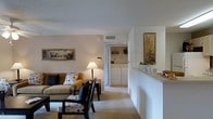 This single-floor residence offers a thoughtfully designed layout that balances comfort and functionality. The living area serves as the heart of the space, featuring warm neutral tones, plush carpeted flooring, and soft ambient lighting provided by a classic table lamp. Framed artwork adorns the walls, adding character and warmth to the room. Seating is arranged around a dark wood coffee table, creating an inviting area for relaxation or conversation.

Adjacent to the living area, a raised counter seamlessly connects to the kitchen space, providing a casual dining or serving surface. A decorative bowl of fresh fruit and kitchen accessories are visible atop the counter, hinting at a practical and welcoming kitchen nearby.

One of the standout features of this residence is the in-unit laundry area, visible through a doorway directly from the living room. The dedicated laundry space houses a full-size washer and dryer along with built-in shelving for storage, offering exceptional convenience.

The overall ambiance of the space is warm and residential, with consistent neutral wall tones and carpeting throughout that create a cohesive, comfortable feel. The open sightlines between rooms contribute to a sense of spaciousness, making this a highly functional and livable single-floor home.