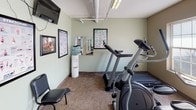 This fitness center is a compact yet comprehensively outfitted single-floor exercise facility designed to support a full range of workout routines. The space features soft sage-green walls, durable carpet tile flooring, and a combination of natural and overhead fluorescent lighting that keeps the environment bright and functional throughout the day. Large windows along one wall invite natural light and offer open outdoor views, creating an airy atmosphere that enhances the workout experience.

The facility is equipped with a variety of cardio machines, including a treadmill, elliptical trainer, and stationary bike, providing options for users of all fitness levels. A multi-station cable strength machine serves as a centerpiece for resistance training, while a dedicated rack holds an assortment of dumbbells for free weight exercises. Suspension training straps and resistance bands are mounted to the wall, offering additional versatility for bodyweight and functional fitness routines.

A wall-mounted flat-screen television provides entertainment during workouts, while framed stretching and exercise reference charts are displayed throughout the space for guidance. A water cooler ensures convenient hydration, and seating is available along the wall for rest between sets. The space is thoughtfully organized to maximize usability while maintaining an open, accessible layout suitable for a shared fitness environment.