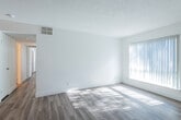 This single-story space features a cohesive, light-filled design unified by wood-style laminate flooring and crisp white walls throughout. The living area is generously proportioned, bathed in natural light streaming through large windows fitted with vertical blinds. A textured popcorn ceiling and integrated HVAC vents contribute to the room's functional layout, while a ceiling fan adds comfort. The space flows openly toward the front entry, where a stainless steel refrigerator and track lighting mark the transition into the kitchen.

The kitchen is efficiently designed with dark laminate countertops, white cabinetry, a stainless steel undermount sink with a pull-down faucet, and built-in appliances. Upper shelving with warm wood accents adds a subtle contrast to the otherwise clean aesthetic.

The bedrooms are well-sized, each receiving ample natural light through large windows with vertical blinds and equipped with ceiling fans. One bedroom features a walk-in closet alongside an additional standard closet, offering generous storage. A digital thermostat is conveniently mounted in the hallway.

The bathroom maintains the space's bright, clean character with a wide vanity, integrated sink, large mirror, and wood-toned flooring, completing a well-appointed and cohesive interior.