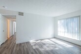 This single-story space features a cohesive, light-filled design unified by wood-style laminate flooring and crisp white walls throughout. The living area is generously proportioned, bathed in natural light streaming through large windows fitted with vertical blinds. A textured popcorn ceiling and integrated HVAC vents contribute to the room's functional layout, while a ceiling fan adds comfort. The space flows openly toward the front entry, where a stainless steel refrigerator and track lighting mark the transition into the kitchen.

The kitchen is efficiently designed with dark laminate countertops, white cabinetry, a stainless steel undermount sink with a pull-down faucet, and built-in appliances. Upper shelving with warm wood accents adds a subtle contrast to the otherwise clean aesthetic.

The bedrooms are well-sized, each receiving ample natural light through large windows with vertical blinds and equipped with ceiling fans. One bedroom features a walk-in closet alongside an additional standard closet, offering generous storage. A digital thermostat is conveniently mounted in the hallway.

The bathroom maintains the space's bright, clean character with a wide vanity, integrated sink, large mirror, and wood-toned flooring, completing a well-appointed and cohesive interior.