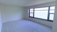 This single-floor space features a generously sized open room finished with light-colored wall-to-wall carpeting that contributes to a clean, neutral aesthetic throughout. The room is defined by a large multi-pane window along one wall, which allows an abundance of natural light to fill the interior while framing views of the surrounding urban cityscape, including neighboring buildings visible from what appears to be an elevated vantage point. The walls are finished in a crisp white tone, complementing the bright atmosphere created by the expansive window. The textured ceiling adds a subtle architectural detail to the otherwise streamlined interior. The open layout of the room offers considerable flexibility for a variety of furniture arrangements and uses. The overall ambiance is bright, airy, and clean, with the urban views serving as a notable visual feature. The simplicity of the finishes and the openness of the floor plan create a versatile and functional environment suited for a range of purposes.