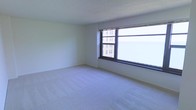 This single-floor space features a generously sized open room finished with light-colored wall-to-wall carpeting that contributes to a clean, neutral aesthetic throughout. The room is defined by a large multi-pane window along one wall, which allows an abundance of natural light to fill the interior while framing views of the surrounding urban cityscape, including neighboring buildings visible from what appears to be an elevated vantage point. The walls are finished in a crisp white tone, complementing the bright atmosphere created by the expansive window. The textured ceiling adds a subtle architectural detail to the otherwise streamlined interior. The open layout of the room offers considerable flexibility for a variety of furniture arrangements and uses. The overall ambiance is bright, airy, and clean, with the urban views serving as a notable visual feature. The simplicity of the finishes and the openness of the floor plan create a versatile and functional environment suited for a range of purposes.