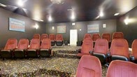Located at 1025 Kensington Dr in Jacksonville, NC, this single-floor space is centered around a dedicated private screening room that delivers an authentic cinematic experience. The theater is designed with tiered stadium-style seating, accommodating multiple rows of plush, red upholstered theater chairs with black armrests and built-in cupholders — offering both comfort and a true movie-house feel. The floor is covered in a bold, patterned carpet characteristic of classic cinema interiors, adding a lively visual touch to the otherwise darkened environment. The walls are finished in deep charcoal tones, enhancing the immersive, light-controlled atmosphere ideal for screening. Decorative cinema-themed artwork adorns the walls, reinforcing the entertainment-focused character of the space. Recessed and wall-mounted lighting provides subtle illumination throughout, while built-in speakers support quality audio. A storage cabinet is positioned at the rear of the room, offering practical utility without disrupting the viewing environment. The elevated rear rows ensure unobstructed sightlines for all occupants, making this space well-suited for private film screenings, presentations, or group entertainment gatherings.