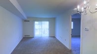 This single-floor space features an open and functional layout designed to maximize usability. The main living area is finished with wall-to-wall carpeting in a neutral tone, providing a comfortable and cohesive foundation throughout. A large sliding glass door serves as a focal point of the room, fitted with vertical blinds that allow for adjustable light control while offering a view of the outdoors. The door also provides convenient access to an exterior space. Natural light flows generously into the interior through this expansive glazed opening, contributing to a bright and airy atmosphere. The walls are finished in a clean white, enhancing the sense of openness throughout the space. HVAC vents are positioned both on the wall and along the baseboard, ensuring climate control across the area. An open doorway on the right side of the main living area connects to an additional room, also carpeted, suggesting a versatile layout suited for multiple living configurations. The overall design is straightforward and practical, with a neutral palette that provides flexibility for a variety of interior arrangements and personal styles.