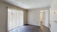 This single-floor residence offers a practical and comfortable layout with consistent dark hardwood-style flooring throughout. The main living area is a spacious, open room highlighted by a large sliding glass door fitted with vertical blinds, allowing abundant natural light to fill the space. Neutral beige walls and a textured popcorn ceiling complete the room's clean, understated aesthetic.

The galley-style kitchen is efficiently designed with rich dark cabinetry lining both sides, light granite-style countertops, and black appliances including a range, dishwasher, and refrigerator. A ceiling-mounted light fixture provides bright, functional illumination, and a door at the end of the kitchen offers additional access within the unit.

The bedroom features dark hardwood-style flooring, a ceiling fan with a light fixture, and a window with exterior views, providing natural ventilation and light. The bedroom connects directly to a walk-in closet and a full bathroom, which includes a tub-shower combination with tile surround, a toilet, and a vanity with dark cabinetry. The layout promotes a sense of privacy and convenience within the bedroom suite.

Overall, the space presents a cohesive design with neutral tones, durable flooring, and functional room arrangements suited for everyday living.