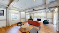 This well-appointed clubhouse in Columbia, SC offers a welcoming and functional gathering space designed with both comfort and style in mind. The open-plan layout is anchored by warm hardwood flooring that runs throughout, complemented by a striking coffered ceiling with exposed beams, recessed lighting, and a ceiling fan that enhance the architectural character of the space.

The lounge area serves as the heart of the clubhouse, centered around a floor-to-ceiling white painted brick fireplace that provides a bold focal point. Comfortable mid-century modern seating — including a rich terracotta sofa, teal loveseats, and accent chairs — is arranged around a woven ottoman and a wood coffee table atop a colorful area rug, creating an inviting atmosphere for socializing. Large sliding glass doors and windows along multiple walls flood the space with natural light while offering views of the surrounding outdoor amenities, including what appears to be a pool area.

The kitchen area is equally impressive, featuring a large light blue island with a dramatic marble-look countertop and industrial-style bar stools. Stainless steel appliances, open shelving, and a decorative patterned backsplash round out this well-equipped space, making it ideal for hosting events and gatherings. The cohesive design throughout reflects a modern yet comfortable clubhouse environment.