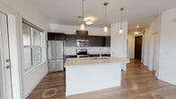 This single-floor unit presents a clean, contemporary design characterized by an open-concept layout that seamlessly connects the kitchen and living area. The kitchen is a standout feature, equipped with stainless steel appliances including a refrigerator, electric range, and built-in microwave. Dark espresso cabinetry extends to the ceiling, providing ample storage, while a large white island with a built-in sink and light quartz countertops serves as both a functional workspace and a natural gathering point. Pendant lighting overhead adds a polished finishing touch. Wood-look flooring runs throughout the main living areas, contributing to a warm yet modern aesthetic, while the open living space offers generous room for furnishings and everyday living.

The bedroom features neutral carpet flooring and a broad window that invites natural light and provides exterior views. The bathroom is well-appointed with wood-look tile flooring, a dark vanity with an integrated sink, and a full soaking tub framed by neutral tile surround. The overall color palette—white walls, dark cabinetry, and light countertops—creates a cohesive, move-in ready interior with a contemporary feel throughout.
