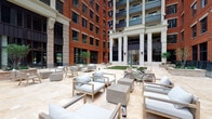 Located at 205 S High St in the heart of Columbus, Ohio, this outdoor courtyard space offers a refined urban retreat within a distinguished mixed-use development. The courtyard is framed by soaring multi-story brick facades accented with large industrial-style windows, white classical columns, and decorative wall sconces that lend a blend of historic character and contemporary elegance.

The ground-level patio is finished in light travertine pavers, creating a clean and sophisticated surface that extends throughout the seating areas. Generously spaced groupings of modern wood-framed lounge chairs with thick grey cushions and concrete coffee tables are arranged to encourage both conversation and relaxation. A patch of artificial turf adds a touch of greenery to the hardscape environment, complementing the potted trees and plantings that soften the urban setting.

The open-air design allows for abundant natural light and open sky views, making the courtyard an inviting space throughout the day. Ambient string lighting suggests the area transitions seamlessly into an equally welcoming evening setting. Bicycle parking within the courtyard reflects a pedestrian-friendly, community-oriented atmosphere. Overall, the space strikes a balance between architectural grandeur and approachable, comfortable outdoor living in a vibrant downtown Columbus location.