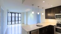 This single-floor space showcases a clean, contemporary design centered around an open-concept layout that seamlessly connects the kitchen and living areas. The kitchen is appointed with sleek dark cabinetry that creates a striking contrast against the bright white countertops, which extend into a spacious island/peninsula with an undermount stainless steel sink. Three stylish pendant lights hang above the counter space, providing focused task lighting while enhancing the modern aesthetic. Recessed lighting and a track lighting fixture further illuminate the space throughout. The living area flows naturally from the kitchen, benefiting from large floor-to-ceiling windows along one wall that invite abundant natural light and frame views of the neighboring urban architecture. The light-toned flooring throughout reinforces the bright, airy feel of the interior. A separate alcove or additional room is visible through an open doorway, offering functional versatility. The overall ambiance is polished and minimalist, with a neutral color palette and thoughtful design details that create a cohesive, modern living environment.