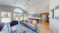 Located at 1421 Carolina Ct in Schaumburg, IL, this single-floor community clubhouse delivers a sophisticated and welcoming atmosphere through its thoughtfully designed open-concept layout. The space is anchored by dramatic vaulted ceilings that soar overhead, amplifying the sense of openness throughout. A striking arched window wall floods the interior with abundant natural light while framing sweeping views of the outdoor pool and patio area, seamlessly connecting the indoors with the outdoors.

The lounge area is generously appointed with plush seating, including a tufted bench, sectional sofas, and accent chairs in coordinating navy and yellow tones, all set atop richly patterned area rugs over light hardwood flooring. A tile-clad fireplace wall with a large wall-mounted television anchors a separate seating nook, creating an inviting space for relaxation or casual gatherings.

The full kitchen is equipped with stainless steel appliances, warm wood cabinetry with gold hardware, a deep double sink, a dishwasher, and an expansive white quartz island with bar-height seating. Floating shelves on a slate-blue accent wall add both style and function. A built-in beverage station further enhances the space's entertaining capabilities. Track lighting and pendant fixtures illuminate the entire space with precision and warmth.