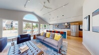 Located at 1421 Carolina Ct in Schaumburg, IL, this single-floor community clubhouse delivers a sophisticated and welcoming atmosphere through its thoughtfully designed open-concept layout. The space is anchored by dramatic vaulted ceilings that soar overhead, amplifying the sense of openness throughout. A striking arched window wall floods the interior with abundant natural light while framing sweeping views of the outdoor pool and patio area, seamlessly connecting the indoors with the outdoors.

The lounge area is generously appointed with plush seating, including a tufted bench, sectional sofas, and accent chairs in coordinating navy and yellow tones, all set atop richly patterned area rugs over light hardwood flooring. A tile-clad fireplace wall with a large wall-mounted television anchors a separate seating nook, creating an inviting space for relaxation or casual gatherings.

The full kitchen is equipped with stainless steel appliances, warm wood cabinetry with gold hardware, a deep double sink, a dishwasher, and an expansive white quartz island with bar-height seating. Floating shelves on a slate-blue accent wall add both style and function. A built-in beverage station further enhances the space's entertaining capabilities. Track lighting and pendant fixtures illuminate the entire space with precision and warmth.