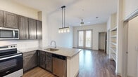 This single-story unit in Austin, Texas offers a thoughtfully designed open-concept layout that seamlessly connects the kitchen and living areas. The kitchen serves as a focal point, showcasing dark espresso cabinetry, granite countertops, and a peninsula with an undermount sink and pendant lighting overhead. Stainless steel appliances, including a refrigerator, electric range, and built-in microwave, complete the space. Wood-style plank flooring runs throughout the kitchen and living areas, while a ceiling fan and glass french doors lead to a private balcony, allowing natural light to fill the space and providing outdoor views.

The bedroom features plush carpeting, a large window with outdoor views, and direct access to a walk-in closet outfitted with multi-tier wire shelving for organized storage. A separate closet provides additional convenience. The neutral color palette throughout creates a clean, modern ambiance. The unit's efficient floor plan maximizes usable space, making it a practical and comfortable home in the 78741 zip code of Austin.