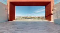 This commanding elevated terrace makes an immediate architectural statement through its bold, oversized red-rendered frame — a defining structural element that draws the eye outward toward an expansive panoramic cityscape. The terrace floor is finished in large-format grey stone tiles, laid in a clean grid pattern that reinforces the space's sense of order and scale, stretching generously from the interior threshold to an open-air vantage point.

A glass balustrade at the terrace's edge preserves unobstructed sightlines while ensuring safety, allowing visitors to take in the full sweep of the urban landscape below. The view encompasses a sprawling city grid, dotted with mid-rise buildings, green tree canopies, and distant high-rise structures, all set against a backdrop of arid mountain ranges under a clear blue sky.

The interplay between the vivid terracotta-red of the architectural surround and the cool grey of the tile flooring creates a visually striking contrast, lending the space a bold, contemporary aesthetic. The open exposure to natural light and sky enhances the sense of openness and grandeur. This terrace functions as both an architectural feature and an observation platform, offering an impressive, uninterrupted connection to the surrounding urban and natural environment.