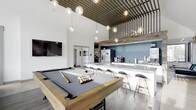 Located at 8115 Worthington Galena Road in Westerville, Ohio, this single-floor amenity space delivers a polished, modern atmosphere designed for socializing and recreation. The open-concept layout is anchored by a prominent billiards area, complete with a wood-framed pool table set atop wide-plank hardwood-style flooring. Soaring ceilings amplify the sense of spaciousness, drawing the eye upward to a striking slatted wood ceiling accent that adds warmth and industrial character to the room. A cascading arrangement of globe pendant lights provides both ambient illumination and a refined visual focal point overhead. Adjacent to the game area, a long white bar counter with bar-height seating offers a casual gathering spot, backed by a teal accent wall and a stainless steel refrigerator, with a beverage and coffee station visible along the counter. A wall-mounted flat-screen television is positioned to serve the lounge area, making this space well-suited for watching events or entertaining. The overall design blends rustic elements — including the reclaimed wood ceiling details — with contemporary finishes, creating an inviting and versatile communal environment.