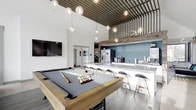 Located at 8115 Worthington Galena Road in Westerville, Ohio, this single-floor amenity space delivers a polished, modern atmosphere designed for socializing and recreation. The open-concept layout is anchored by a prominent billiards area, complete with a wood-framed pool table set atop wide-plank hardwood-style flooring. Soaring ceilings amplify the sense of spaciousness, drawing the eye upward to a striking slatted wood ceiling accent that adds warmth and industrial character to the room. A cascading arrangement of globe pendant lights provides both ambient illumination and a refined visual focal point overhead. Adjacent to the game area, a long white bar counter with bar-height seating offers a casual gathering spot, backed by a teal accent wall and a stainless steel refrigerator, with a beverage and coffee station visible along the counter. A wall-mounted flat-screen television is positioned to serve the lounge area, making this space well-suited for watching events or entertaining. The overall design blends rustic elements — including the reclaimed wood ceiling details — with contemporary finishes, creating an inviting and versatile communal environment.