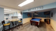 This single-floor communal amenity space is thoughtfully designed to serve residents with a variety of recreational and practical facilities. The interior is unified by a consistent color palette of soft light blue walls paired with white wainscoting, creating a clean and inviting atmosphere throughout.

The billiards area is a standout feature, marked by bold lettered signage and anchored by a full-size pool table with a blue felt surface. A dedicated pendant light hangs above the table, while a wall-mounted fan provides ventilation. A high-top bistro table with bar-height seating sits adjacent, offering a comfortable spot for spectators. Carpet flooring in this zone adds warmth and acoustic comfort.

The laundry facility is well-equipped with multiple rows of front-loading washers and top-loading dryers lining the walls. The machines are neatly arranged and appear coin- or card-operated, providing ample capacity for residents. Light wood-look tile flooring runs throughout this area, keeping the space easy to maintain.

A central entryway connects the zones, featuring a door with direct access to an outdoor stairway, allowing natural light to filter in. Signage indicating a theater and game zone suggests additional dedicated spaces accessible from this common area, rounding out a comprehensive community hub.