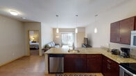 This single-floor residence offers a thoughtfully designed open-concept layout that seamlessly connects the kitchen, dining, and living areas. The kitchen is a standout feature, equipped with rich dark-wood cabinetry, granite countertops, and a full suite of stainless steel appliances including a refrigerator, range, built-in microwave, and dishwasher. A large peninsula with seating for three provides an ideal space for casual dining or entertaining. Three pendant lights hang above the peninsula, adding a stylish focal point to the space. The living area is bright and comfortable, anchored by a sliding glass door that opens to a private balcony, allowing natural light to pour in and offering views of the surrounding neighborhood. The space includes two well-sized bedrooms, each with plush carpeting and ample closet storage, including a walk-in closet in one of the rooms. The full bathroom features hardwood-style flooring, dark-wood vanity cabinetry, a large mirror, and a tub-shower combination. A dedicated in-unit laundry room with a full-size washer and dryer adds everyday convenience. Light hardwood-style flooring flows throughout the main living areas, contributing to a warm and cohesive aesthetic throughout the home.