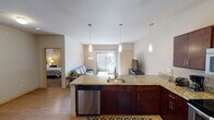 This single-floor residence offers a thoughtfully designed open-concept layout that seamlessly connects the kitchen, dining, and living areas. The kitchen is a standout feature, equipped with rich dark-wood cabinetry, granite countertops, and a full suite of stainless steel appliances including a refrigerator, range, built-in microwave, and dishwasher. A large peninsula with seating for three provides an ideal space for casual dining or entertaining. Three pendant lights hang above the peninsula, adding a stylish focal point to the space. The living area is bright and comfortable, anchored by a sliding glass door that opens to a private balcony, allowing natural light to pour in and offering views of the surrounding neighborhood. The space includes two well-sized bedrooms, each with plush carpeting and ample closet storage, including a walk-in closet in one of the rooms. The full bathroom features hardwood-style flooring, dark-wood vanity cabinetry, a large mirror, and a tub-shower combination. A dedicated in-unit laundry room with a full-size washer and dryer adds everyday convenience. Light hardwood-style flooring flows throughout the main living areas, contributing to a warm and cohesive aesthetic throughout the home.