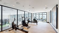 This contemporary fitness center is defined by its striking use of floor-to-ceiling glass walls framed in dark metal, which flood the space with natural light and provide open views of a surrounding courtyard and adjacent building facades. The light hardwood flooring runs throughout, lending warmth to the otherwise clean, minimalist aesthetic characterized by white ceilings and recessed lighting.

The main workout area is well-appointed with cardio equipment, including rowing machines, air bikes, and stationary cycles, all arranged to take full advantage of the panoramic glazing. Large glass partitions divide the fitness zone from neighboring amenity areas, maintaining visual connectivity while defining distinct functional spaces. A glass-paneled entry corridor leads to additional rooms finished with patterned carpet and ambient circular ceiling fixtures, suggesting lounge or meeting areas.

The transparency of the architecture creates a seamless indoor-outdoor feel, with courtyard seating and greenery visible through the expansive windows. Ceiling-mounted HVAC units and integrated speakers reflect a thoughtfully designed infrastructure. The overall ambiance is bright, airy, and professionally curated, making the space well-suited for an active lifestyle within a larger shared amenity environment.