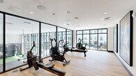 This contemporary fitness center is defined by its striking use of floor-to-ceiling glass walls framed in dark metal, which flood the space with natural light and provide open views of a surrounding courtyard and adjacent building facades. The light hardwood flooring runs throughout, lending warmth to the otherwise clean, minimalist aesthetic characterized by white ceilings and recessed lighting.

The main workout area is well-appointed with cardio equipment, including rowing machines, air bikes, and stationary cycles, all arranged to take full advantage of the panoramic glazing. Large glass partitions divide the fitness zone from neighboring amenity areas, maintaining visual connectivity while defining distinct functional spaces. A glass-paneled entry corridor leads to additional rooms finished with patterned carpet and ambient circular ceiling fixtures, suggesting lounge or meeting areas.

The transparency of the architecture creates a seamless indoor-outdoor feel, with courtyard seating and greenery visible through the expansive windows. Ceiling-mounted HVAC units and integrated speakers reflect a thoughtfully designed infrastructure. The overall ambiance is bright, airy, and professionally curated, making the space well-suited for an active lifestyle within a larger shared amenity environment.