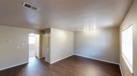 This single-story space presents a straightforward and functional interior characterized by dark hardwood-style flooring that runs throughout, providing a warm contrast to the neutral, light-toned walls. The textured popcorn ceiling adds a classic architectural detail while housing centrally placed HVAC vents for climate control. A track lighting fixture is mounted along the ceiling, offering directed illumination across the main living area.

The open layout allows for versatile furniture arrangement, with the main room connecting seamlessly to an adjacent area visible through an arched doorway. This secondary space benefits from a window that introduces natural light, brightening the transition zone between rooms. An interior door with raised panel detailing and a traditional arched top frame adds a subtle decorative element to the space.

Baseboards run along the perimeter of the walls, and electrical outlets are positioned at standard intervals. The overall ambiance is clean, neutral, and move-in ready, making it well-suited for a variety of uses. The combination of durable flooring, practical lighting, and connected room flow gives the space a balanced and utilitarian character.