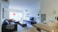 This single-floor space showcases a contemporary open-concept design that seamlessly connects the living and kitchen areas. The living room is anchored by a dark, low-profile media console with floating wall shelves above, providing both functional storage and a clean aesthetic. A flat-screen television is mounted on the console, while decorative accents, including a sculptural horse figurine, add character to the space. The seating area features a dark sectional sofa paired with a modern rectangular coffee table, all set atop a neutral area rug that defines the zone. A large window along the far wall allows generous natural light to fill the room, creating a bright and airy atmosphere. The white walls and light-toned flooring further enhance the sense of openness. Adjacent to the living area, the kitchen features a granite-topped counter with warm golden tones, serving as both a workspace and a casual divider between the two areas. A doorway visible in the background hints at additional rooms beyond, suggesting a well-organized and functional floor plan throughout.