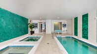 This indoor aquatic facility at 7511 Solstice Cir in Orlando, Florida, offers a well-appointed space designed for relaxation and recreation. The room is anchored by a crystal-clear lap pool and an adjacent hot tub/spa, both finished with vivid blue tile detailing that complements the overall color palette. The walls feature striking panels of hexagonal teal mosaic tiles set against a crisp white backdrop, adding a bold and refreshing visual element to the space. Light-colored, large-format tile flooring with an organic, wave-like pattern extends throughout, contributing to a clean and cohesive aesthetic. Recessed ceiling lighting ensures the space is well-illuminated, while multiple windows along one wall allow natural light to filter in, enhancing the bright and open atmosphere. A dedicated seating area with light aqua-toned chairs and a small side table provides a comfortable spot for relaxation poolside. The space is accessed through glass-panel doors and includes posted spa rules, signage, and safety equipment, reflecting a well-managed and safety-conscious environment. Lush potted plants add a touch of greenery, creating a calm and inviting ambiance throughout this thoughtfully designed indoor aquatic retreat.