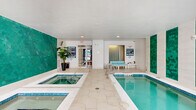 This indoor aquatic facility at 7511 Solstice Cir in Orlando, Florida, offers a well-appointed space designed for relaxation and recreation. The room is anchored by a crystal-clear lap pool and an adjacent hot tub/spa, both finished with vivid blue tile detailing that complements the overall color palette. The walls feature striking panels of hexagonal teal mosaic tiles set against a crisp white backdrop, adding a bold and refreshing visual element to the space. Light-colored, large-format tile flooring with an organic, wave-like pattern extends throughout, contributing to a clean and cohesive aesthetic. Recessed ceiling lighting ensures the space is well-illuminated, while multiple windows along one wall allow natural light to filter in, enhancing the bright and open atmosphere. A dedicated seating area with light aqua-toned chairs and a small side table provides a comfortable spot for relaxation poolside. The space is accessed through glass-panel doors and includes posted spa rules, signage, and safety equipment, reflecting a well-managed and safety-conscious environment. Lush potted plants add a touch of greenery, creating a calm and inviting ambiance throughout this thoughtfully designed indoor aquatic retreat.